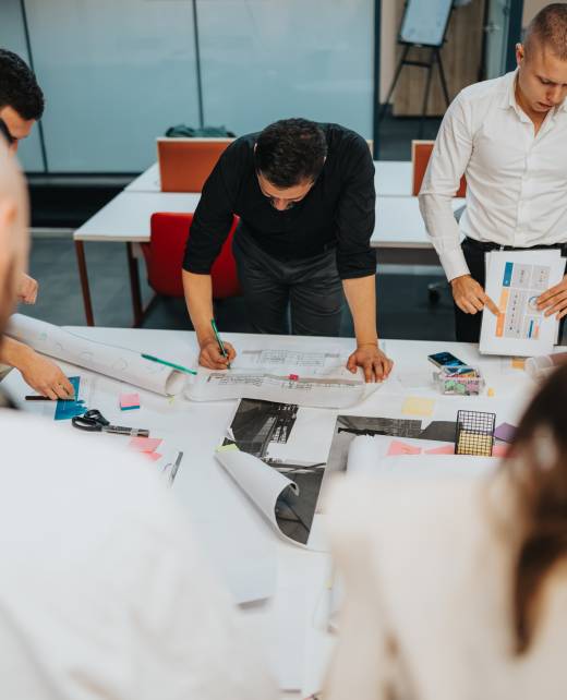 A group of professionals work on architectural plans and sketches around a large table. The scene shows collaboration with papers, diagrams, and planning tools in a modern office.
